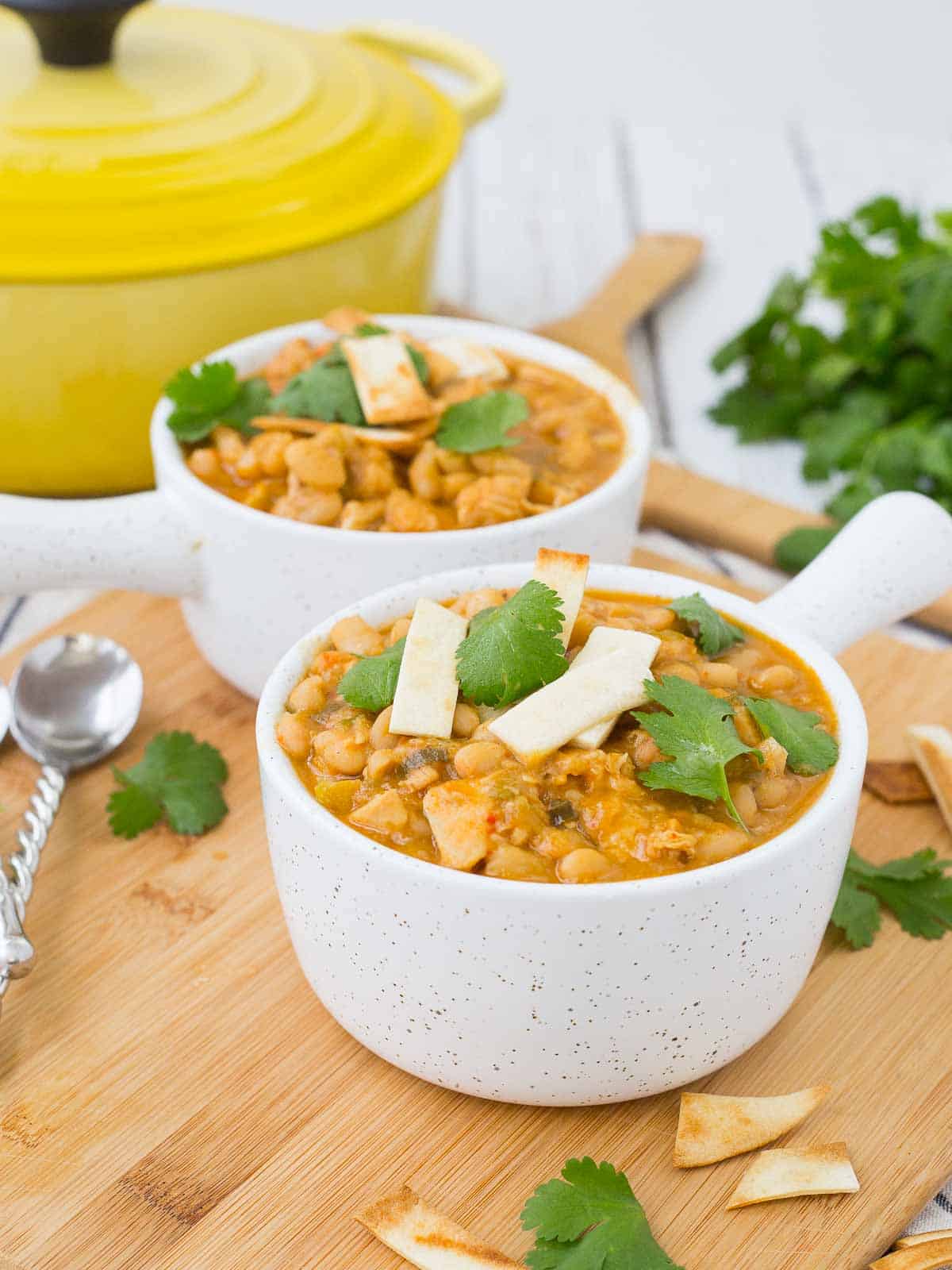 Two servings of chili in white bowls with handle, garnished with parsley leaves and tortilla strips.