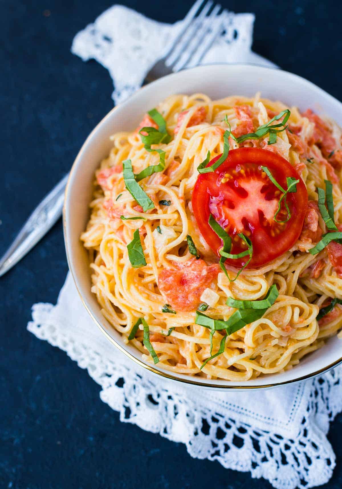 Image of a bowl of angel hair pasta coated in a light cream and fresh tomato sauce. Pasta is garnished with a slice of fresh red tomato and strips of fresh basil. Black background. 