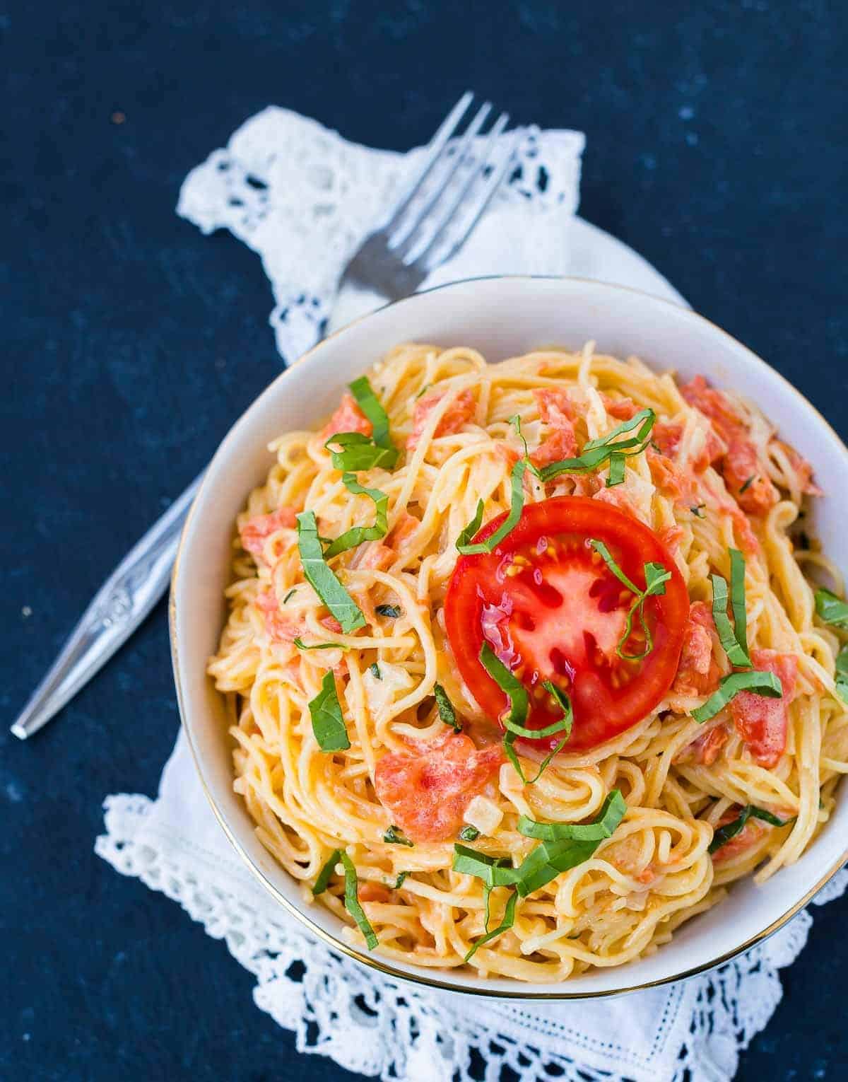 Overhead image of angel hair pasta and a tomato cream sauce. It is garnished with fresh basil and a fresh slice of tomato. A fork and a white linen are also pictured. 
