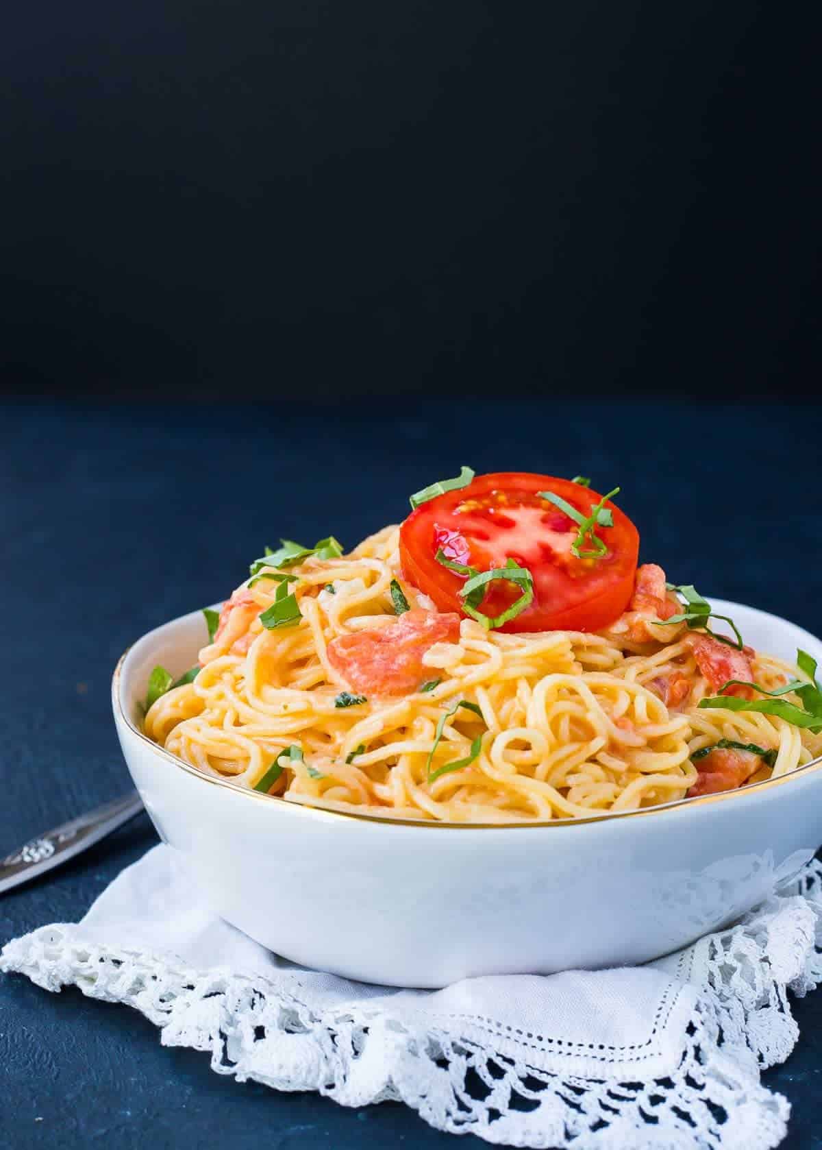 Image of a white bowl on a black background. Bowl is filled with angel hair pasta and a tomato cream sauce. It is garnished with a slice of tomato and fresh basil strips. 