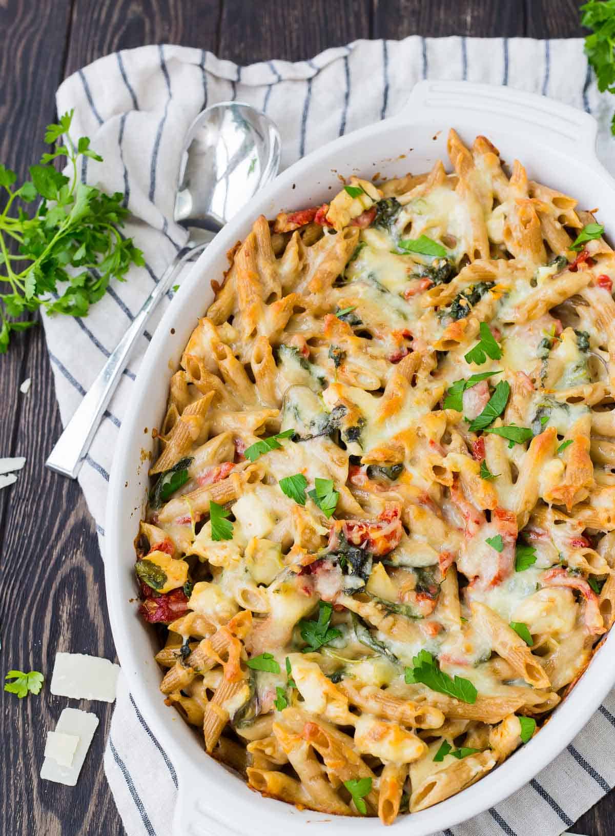 Overhead of baked pasta in white casserole dish, with spoon alongside, on striped cloth.