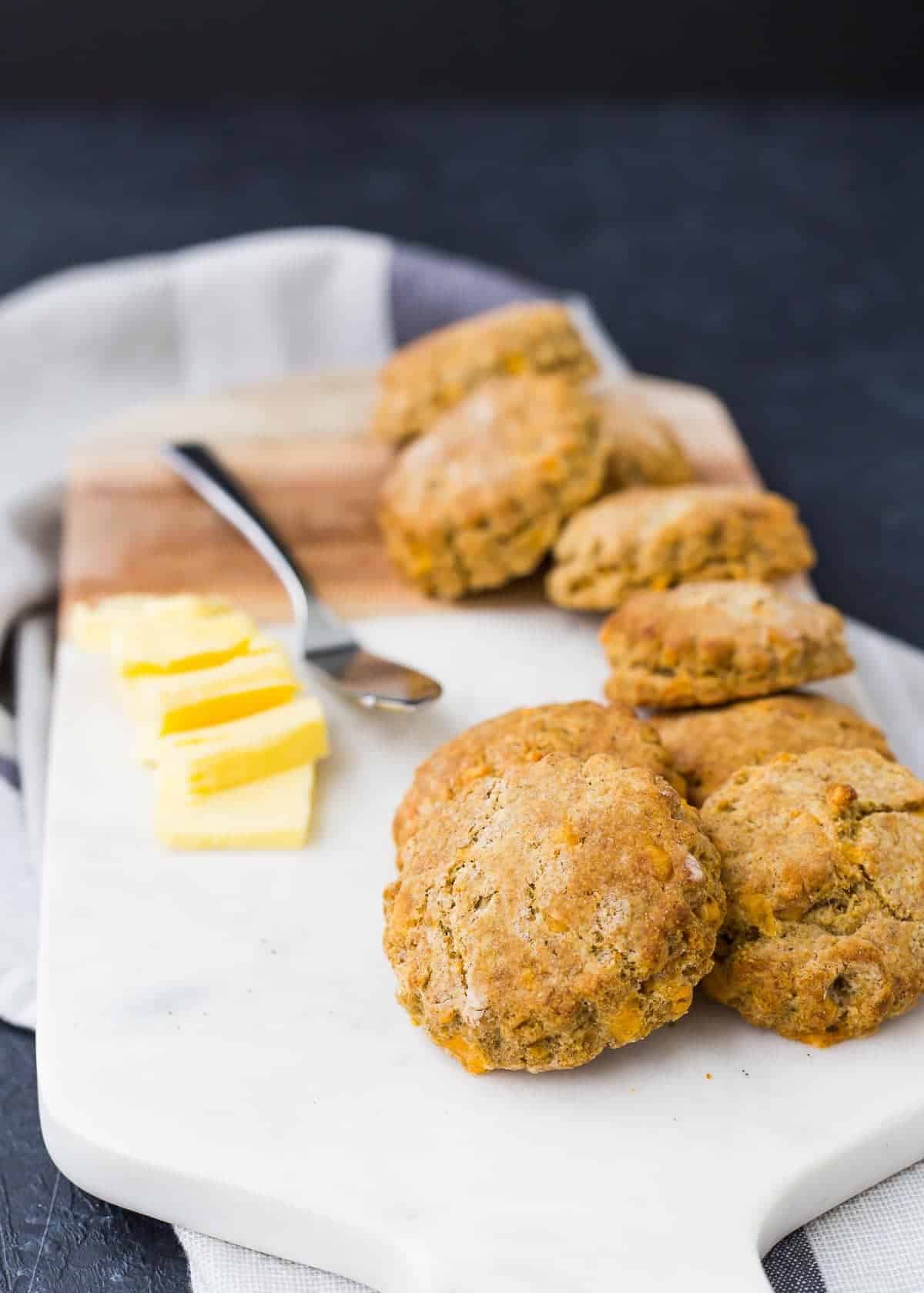 Several biscuits stacked on a white cutting board, with pats of butter and a spreader.