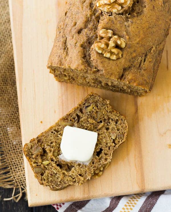 Overhead view of a loaf of apple bread on a wooden cutting board next to a slice topped with butter.