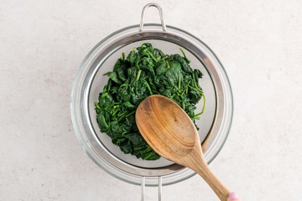 A wooden spoon pressed wilted spinach against a fine mesh strainer to drain the excess liquid.