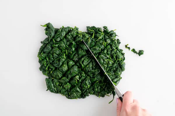 A hand uses a knife to chop cooked spinach on a white surface.