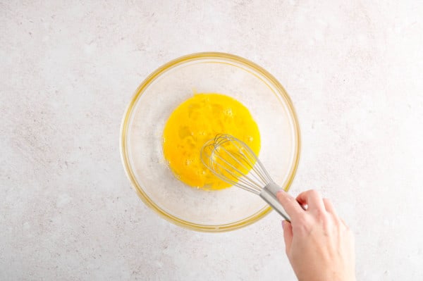 A hand uses a whisk to beat eggs in a glass bowl.