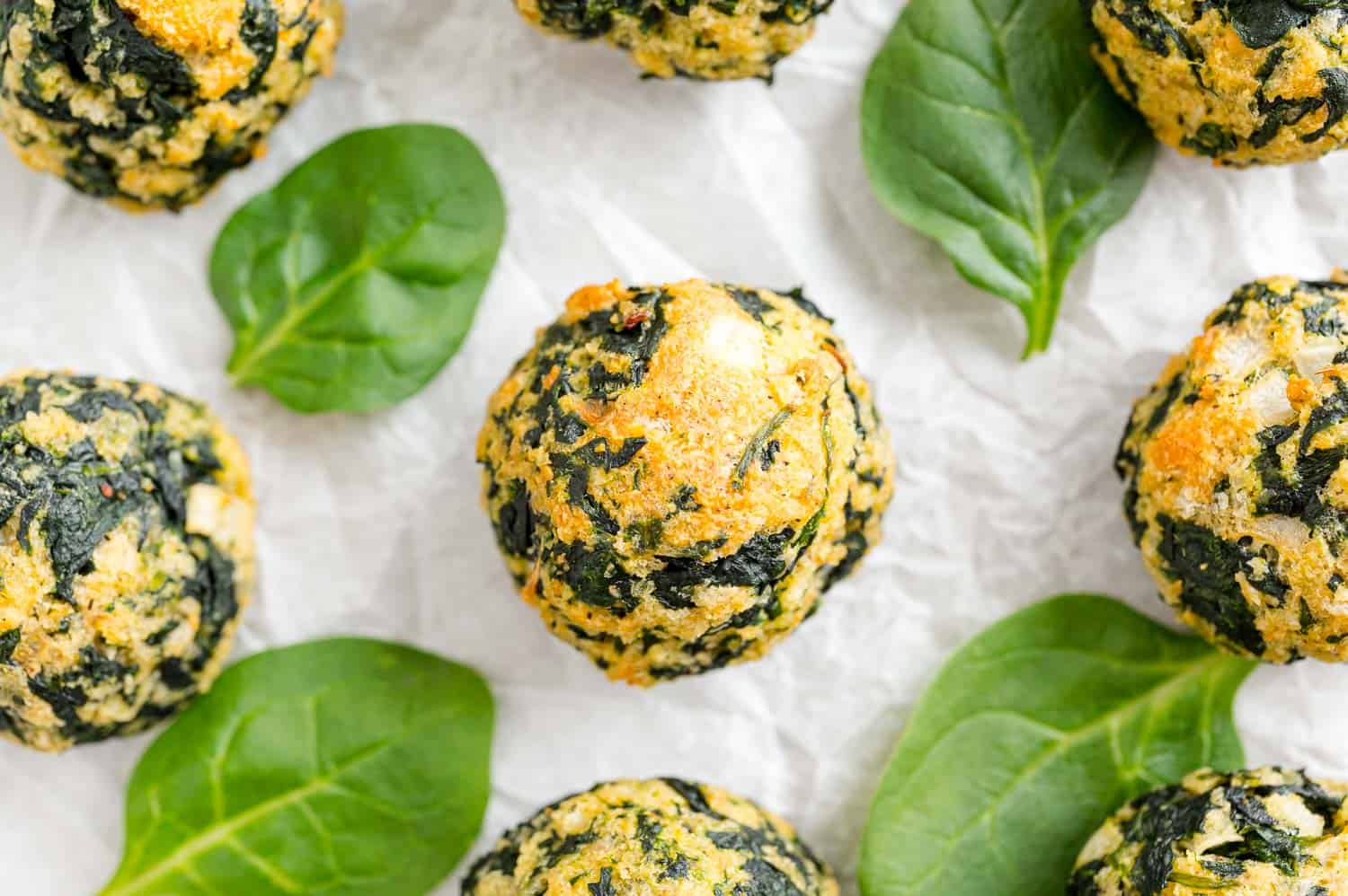 Overhead view of golden baked spinach balls surrounded by spinach leaves on a white surface.