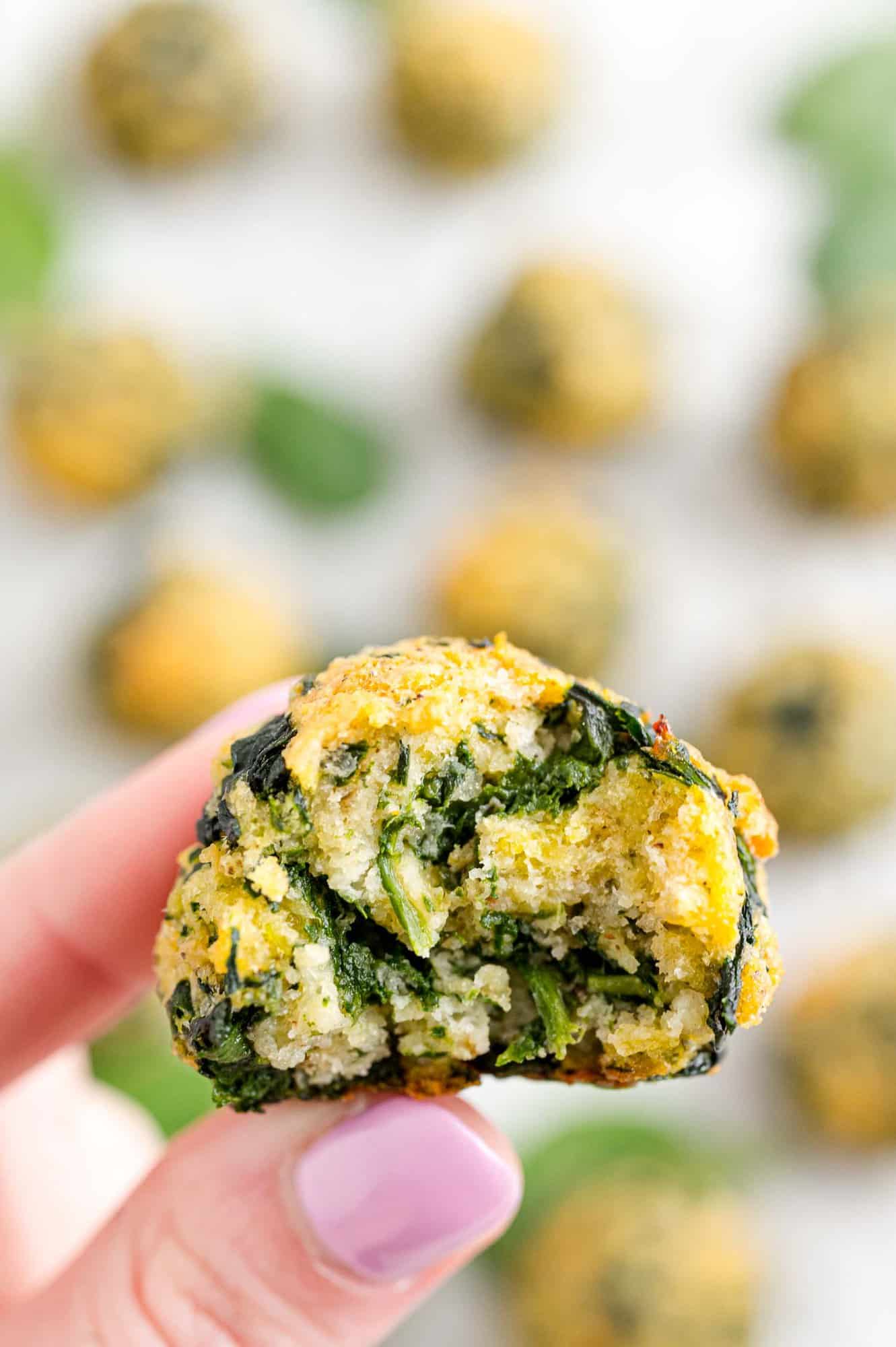 Two fingers holding up a spinach ball with a bite missing, with more spinach balls in the background.