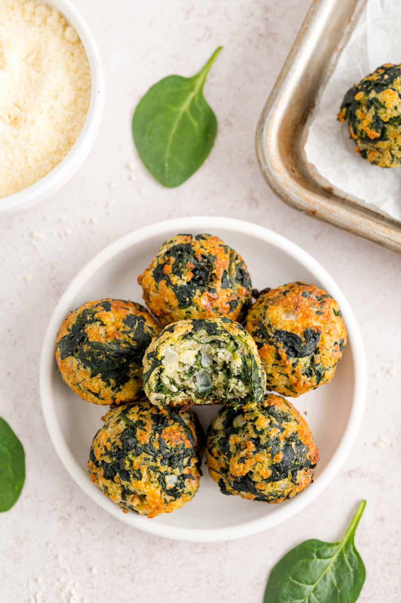 Overhead view of spinach balls stacked on a white plate, with a bite missing from the top spinach ball, next to a bowl of dipping sauce and the corner of a baking sheet.