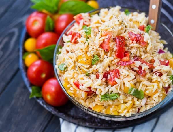 Orzo salad in round bowl, on decorative plate with fresh produce.