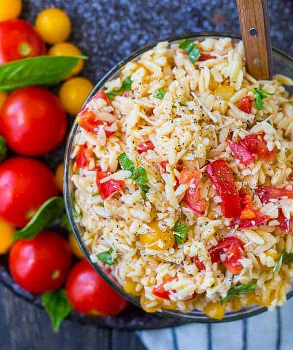 Overhead image of orzo salad in glass bow, with spoon inserted, and fresh whole tomatoes alongside.