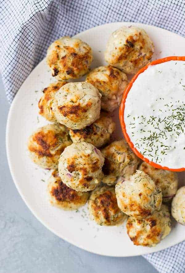 Partial overhead of several meatballs on white plate with orange bowl containing tzatziki sauce.