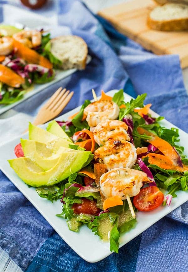 Plated shrimp salad on blue cloth, with sliced bread on wooden cutting board in background.