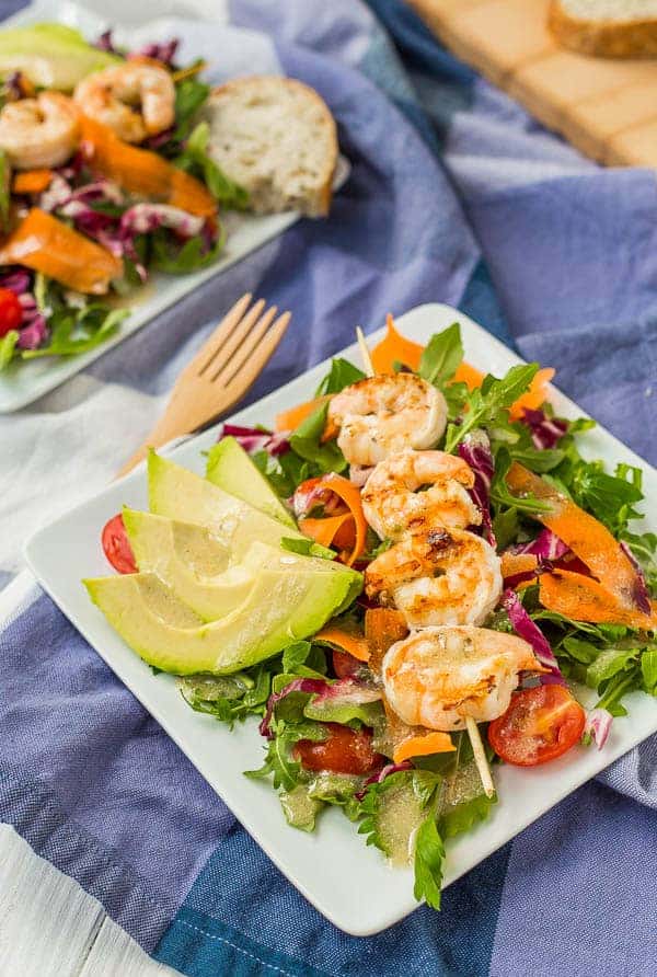 Overhead of grilled shrimp on skewer, plated with salad and wedges of avocado, on square white plate. 