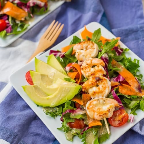 Grilled shrimp on skewer, served on salad and garnished with sliced avocado, on square white plate.