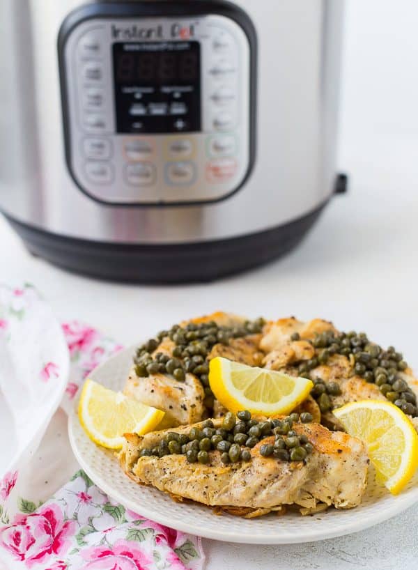 Chicken with capers and lemon slices in front of an instant pot on a white background.