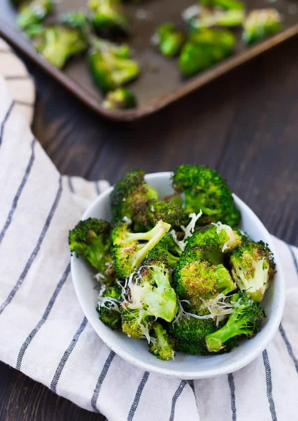 A small white bowl containing roasted broccoli on a white striped dish towel.