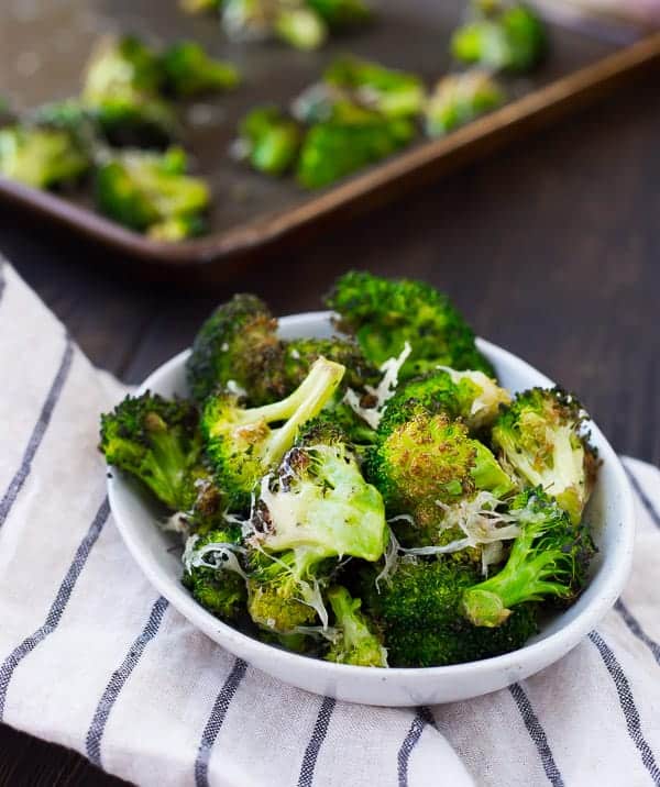 Small white bowl of roasted broccoli on white striped dishtowel, with roasting pan and broccoli in background.