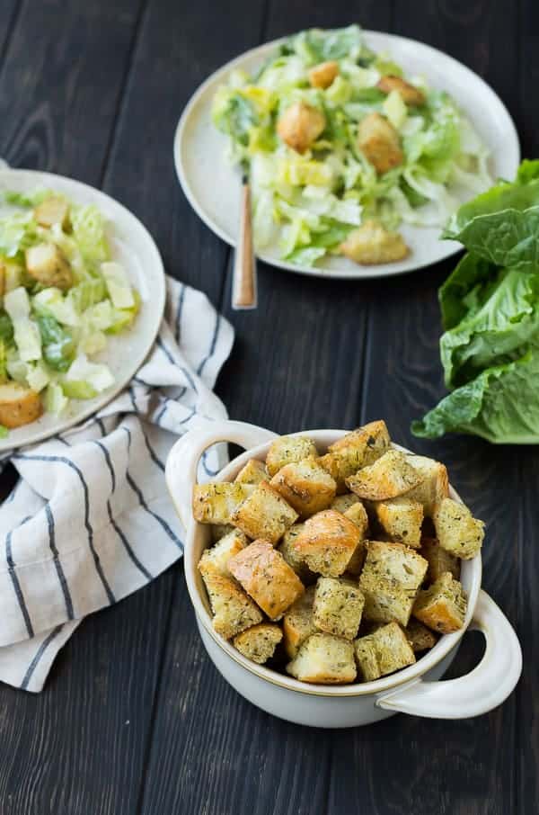 Bowl with Caesar croutons in foreground, two Caesar salads in background.