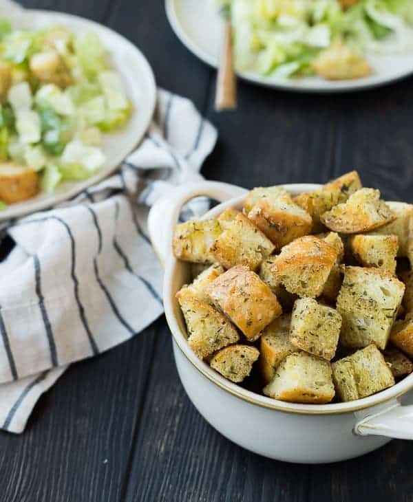 Bowl with Caesar croutons in foreground, two Caesar salads in background.