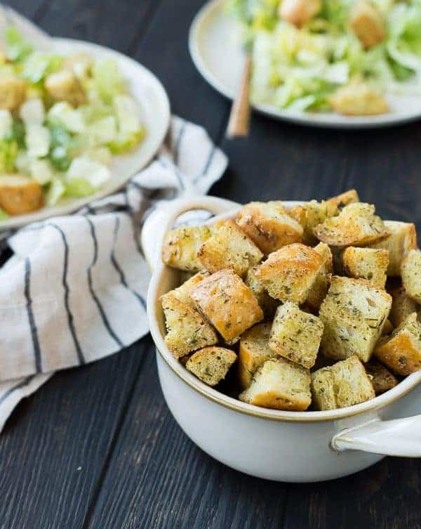 Closeup of Caesar croutons in white bowl, with salads in background.