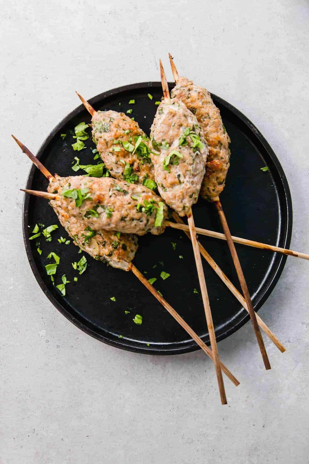 Overhead of several skewered kofta on black round p late, sprinkled with parsley.