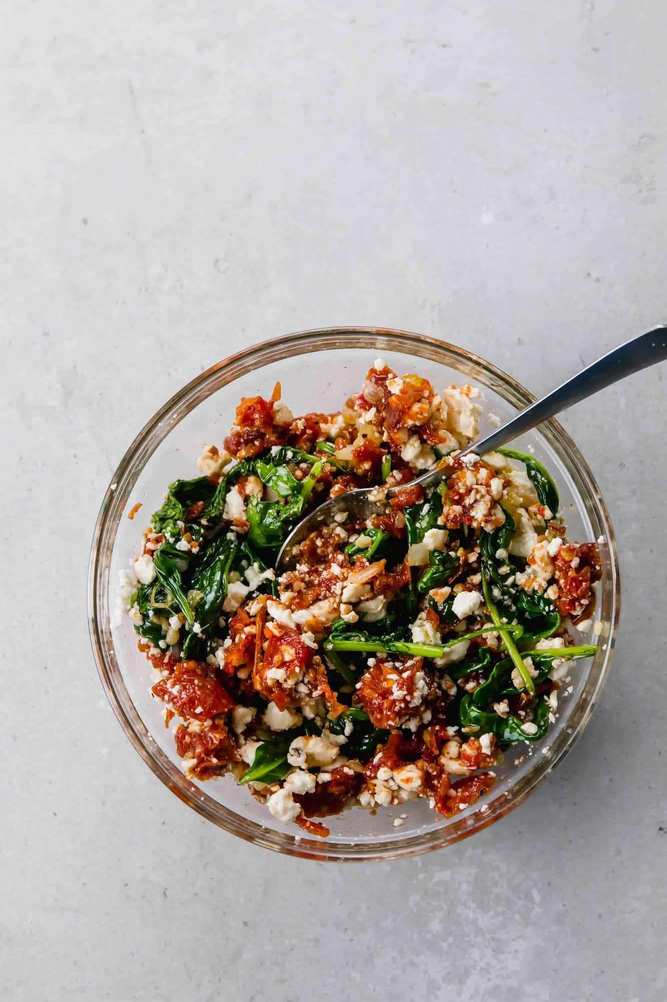Overhead of clear glass bowl containing sundried tomato, kale and feta stuffing, with large spoon.