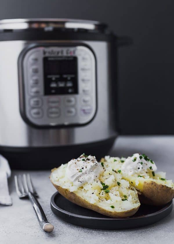 Front view of split baked potato garnished with sour cream and chives, with Instant Pot in background.