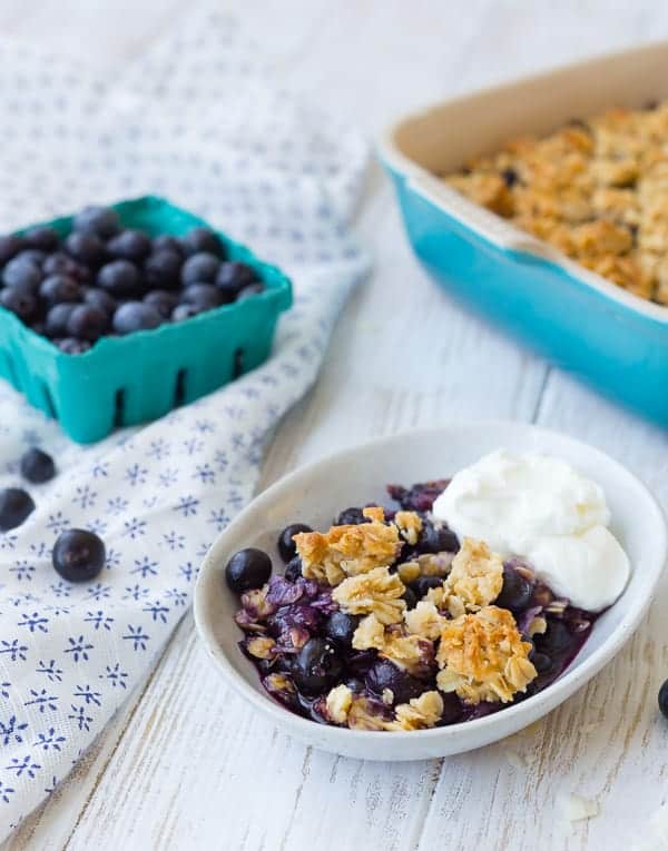 Serving dish containing blueberry crisp with a dollop of yogurt. Fresh blueberries in background.