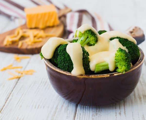 bowl of steamed broccoli topped with cheese sauce. A block of shredded cheddar cheese in background.