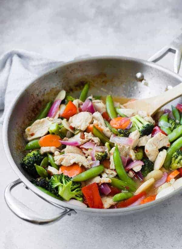 Stainless steel skillet containing chicken stir fry with wooden spoon.