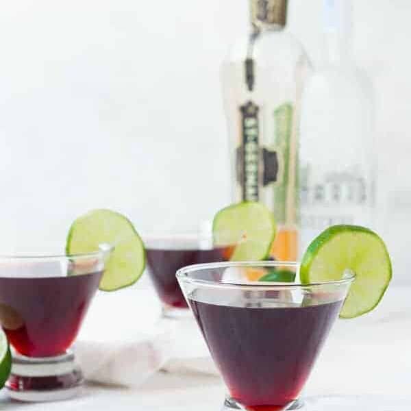 Front view of short martini glasses containing cherry cocktail, with bottles in background.