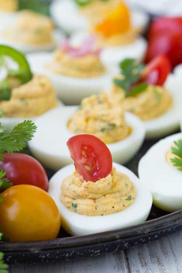 Closeup of a deviled egg topped with a slice of cherry tomato, more tomatoes and eggs in background.