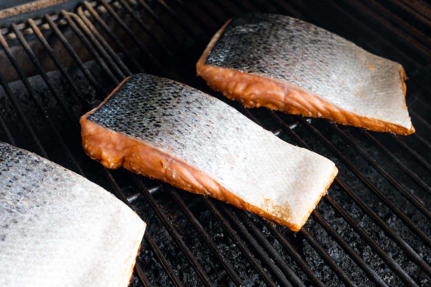 Three salmon fillets grilling skin-side-up on a grill.