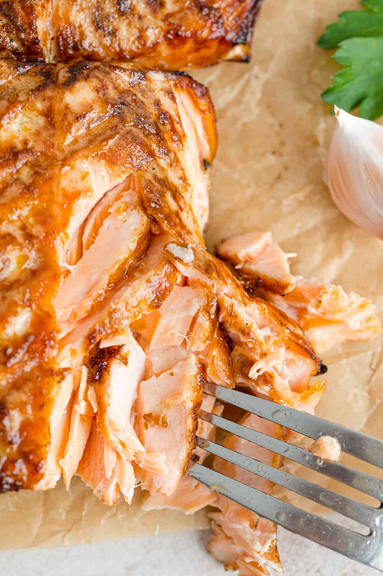 The best grilled salmon recipe for summer and beyond! Enjoy juicy salmon filets marinated in a sweet and garlicky ginger-soy marinade. A fork stuck into flakes of grilled salmon on parchment paper.