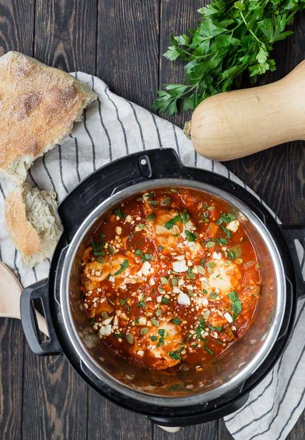 Overhead view of an Instant Pot pressure cooker filled with tomatoes, squash and eggs. Also pictured on a wooden background are a loaf of bread, a whole butternut squash, and fresh parsley.