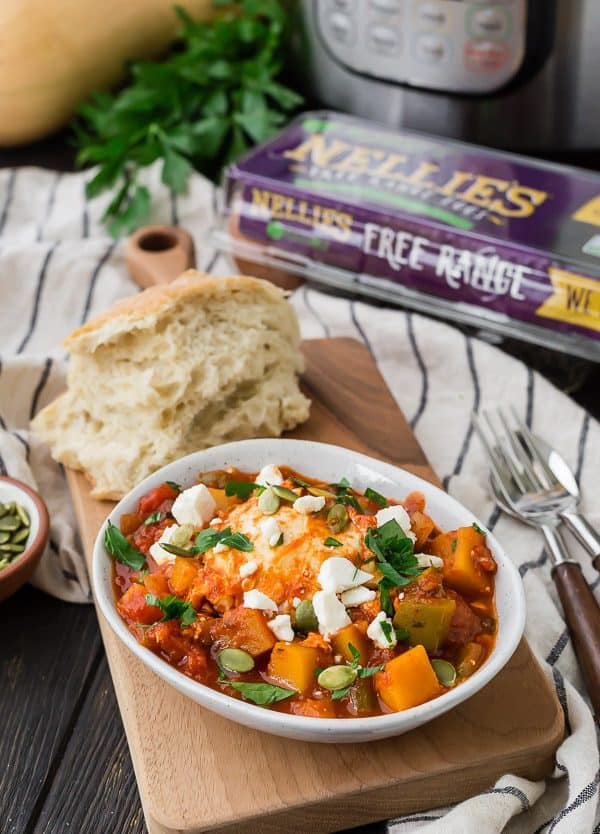Small white bowl filled with butternut squash cubes, tomato sauce, eggs, and feta cheese. In the background, there is bread, a carton of eggs, fresh parsley, and an Instant Pot.
