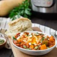 Small white oval bowl filled with tomato sauce, cubed squash, an egg, feta cheese, and pepitas.