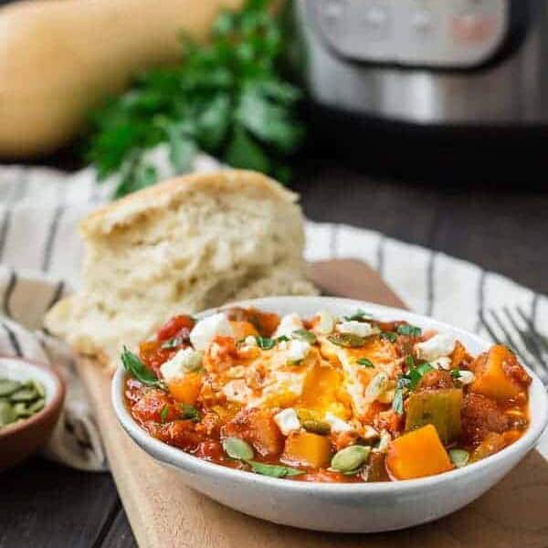 Small white oval bowl filled with tomato sauce, cubed squash, an egg, feta cheese, and pepitas.