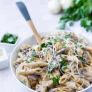 White bowl full of whole wheat penne pasta, chicken, mushrooms, fresh parsley, and parmesan cheese.