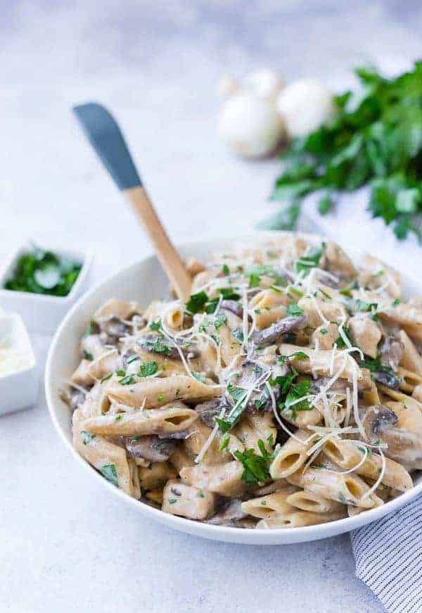 A white bowl full of penne pasta with mushrooms, chicken, parsley, and cheese.