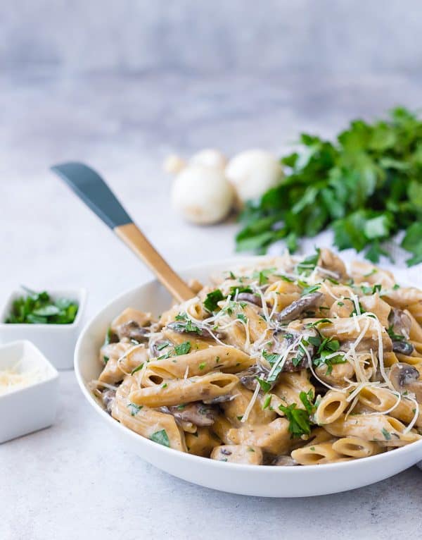 A white bowl filled with whole wheat pasta, mushrooms, and chicken. It is garnished with parsley and cheese.