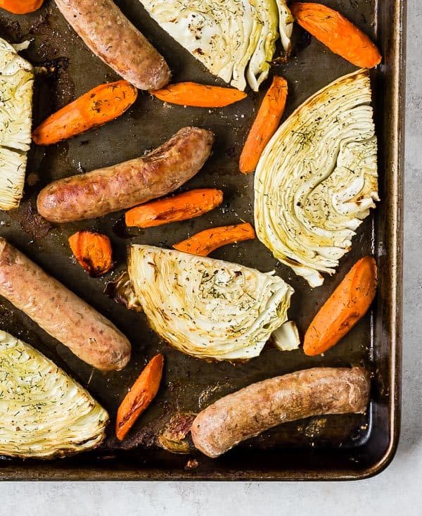 Overhead view of sausages, carrots, and cabbage on a dark gray/brown sheet pan.