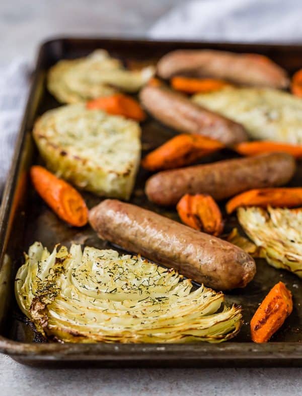 A weathered sheet pan on a gray background, filled with cabbage slices, sausages, and carrots cut on the bias.