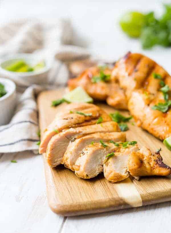 A sliced chicken breast on a cutting board, two other chicken breasts are pictured. They are all sprinkled with fresh cilantro leaves.