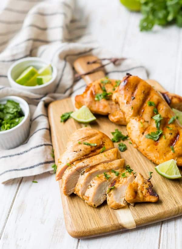 A sliced, grilled chicken breast is on a wooden cutting board with two other chicken breasts. Lime wedges are also pictured.