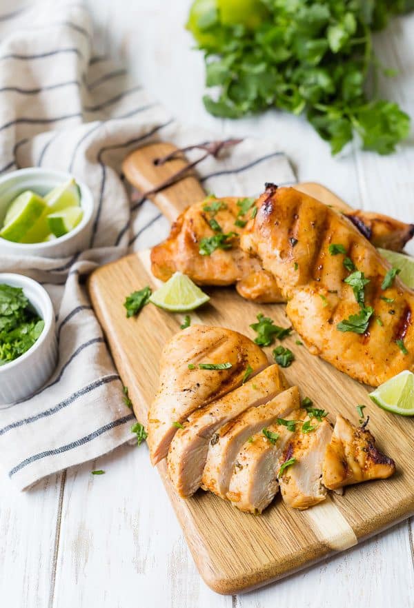 A cutting board with three chicken breasts, one sliced. Lime wedges and fresh cilantro are also pictured.