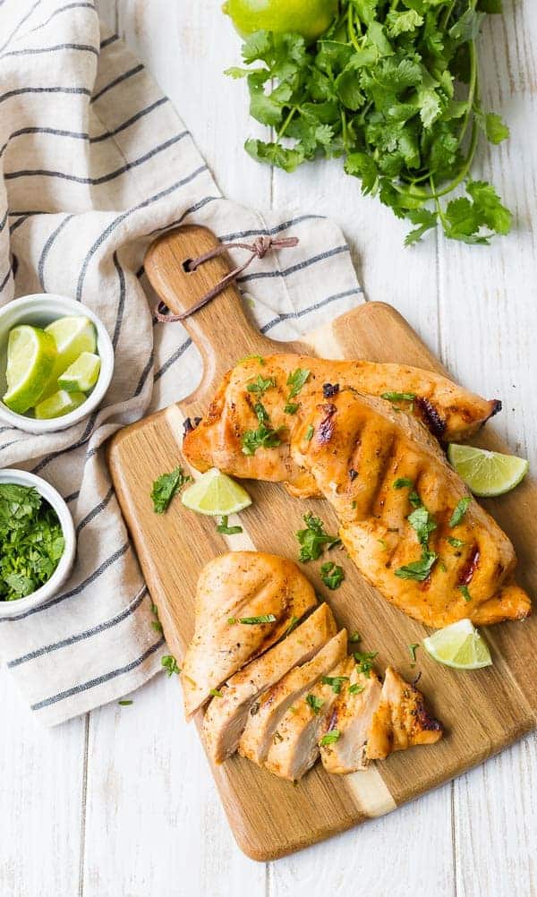 Overhead view of three grilled chicken breasts on a wooden cutting board with a handle. One chicken breast is sliced. All are sprinkled with fresh cilantro.