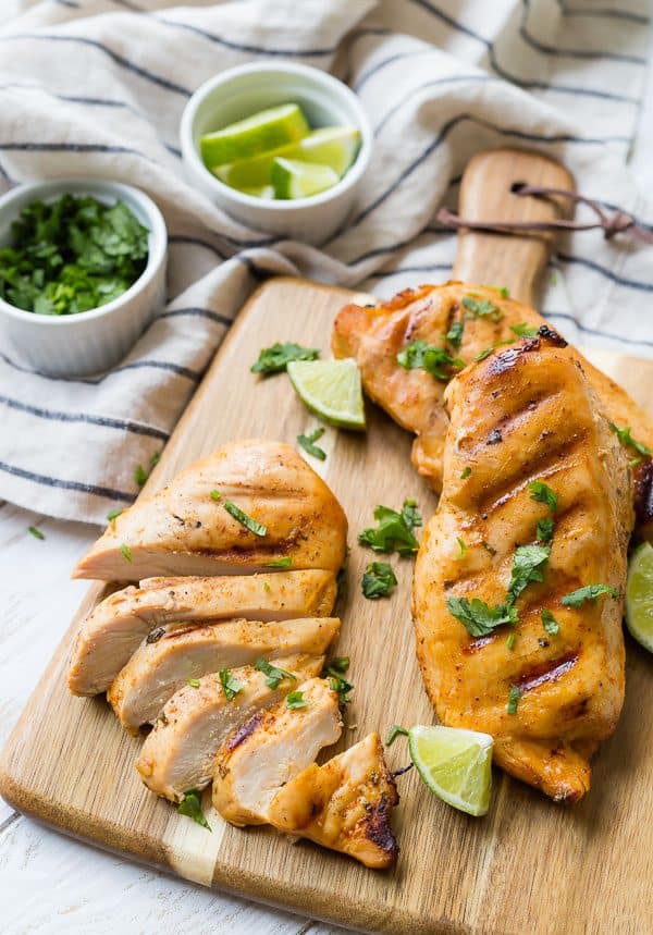 Three chicken breasts, one sliced, on a cutting board with lime and cilantro sprinkled around.