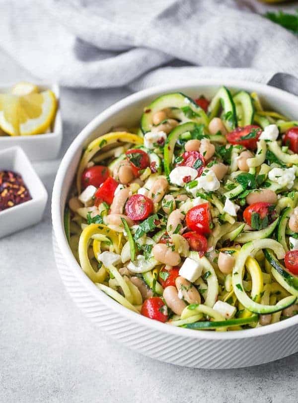 Zucchini noodle salad in white bowl with tomatoes, feta, and beans.